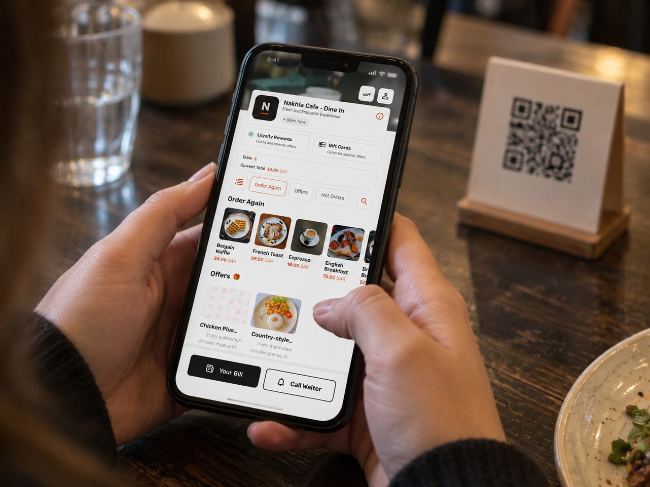 ضيف على طاولة مقهى يحمل هاتفاً يعرض قائمة الصالة، وبطاقة QR الخاصة بالطاولة بجانبه.